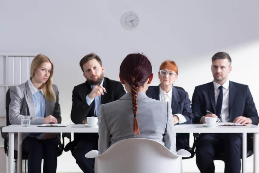 Woman with her back to the camera being interviewed by four serious-looking people