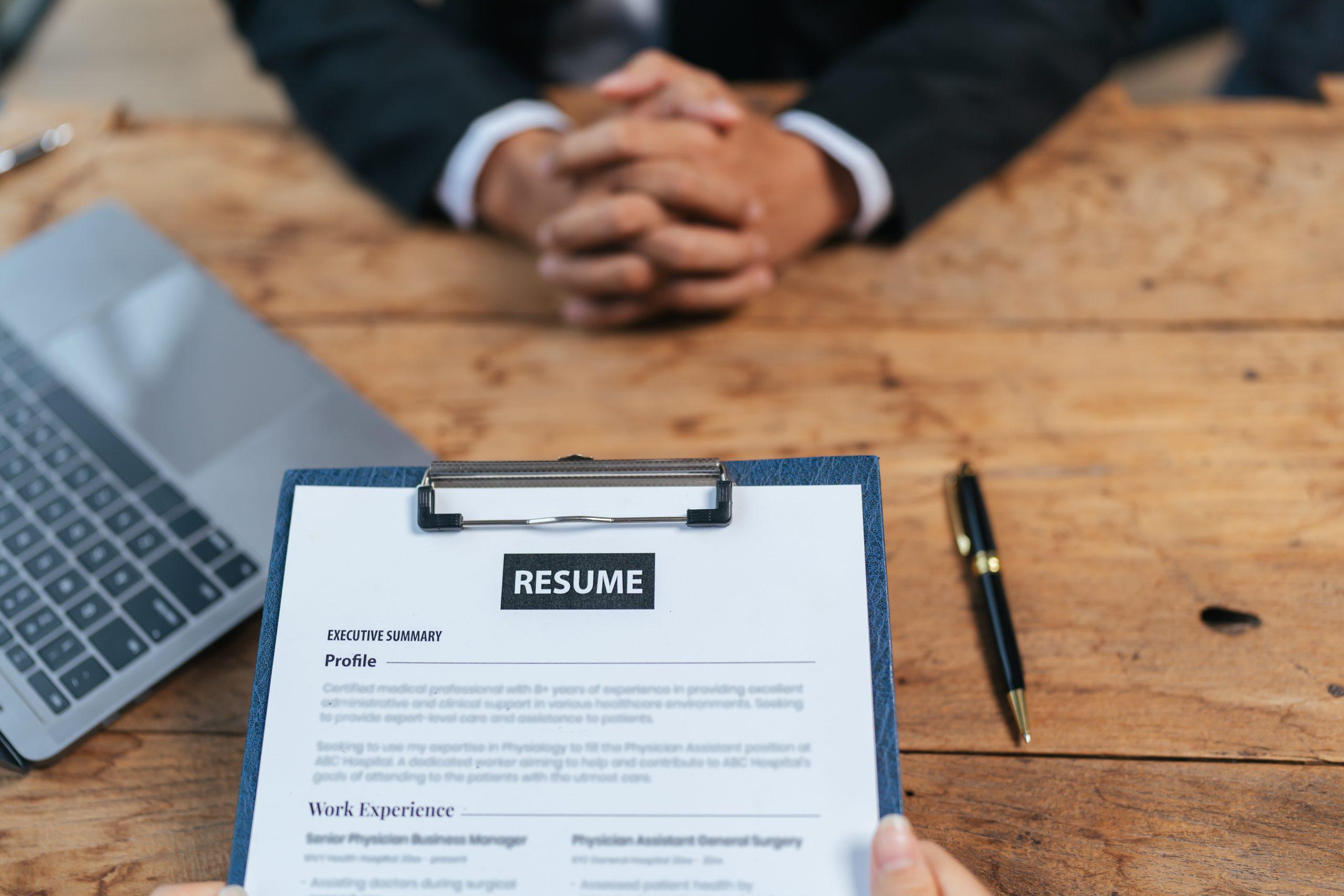 Resume On A Clipboard On A Wooden Table With A Mans Hands Laptop And Pen