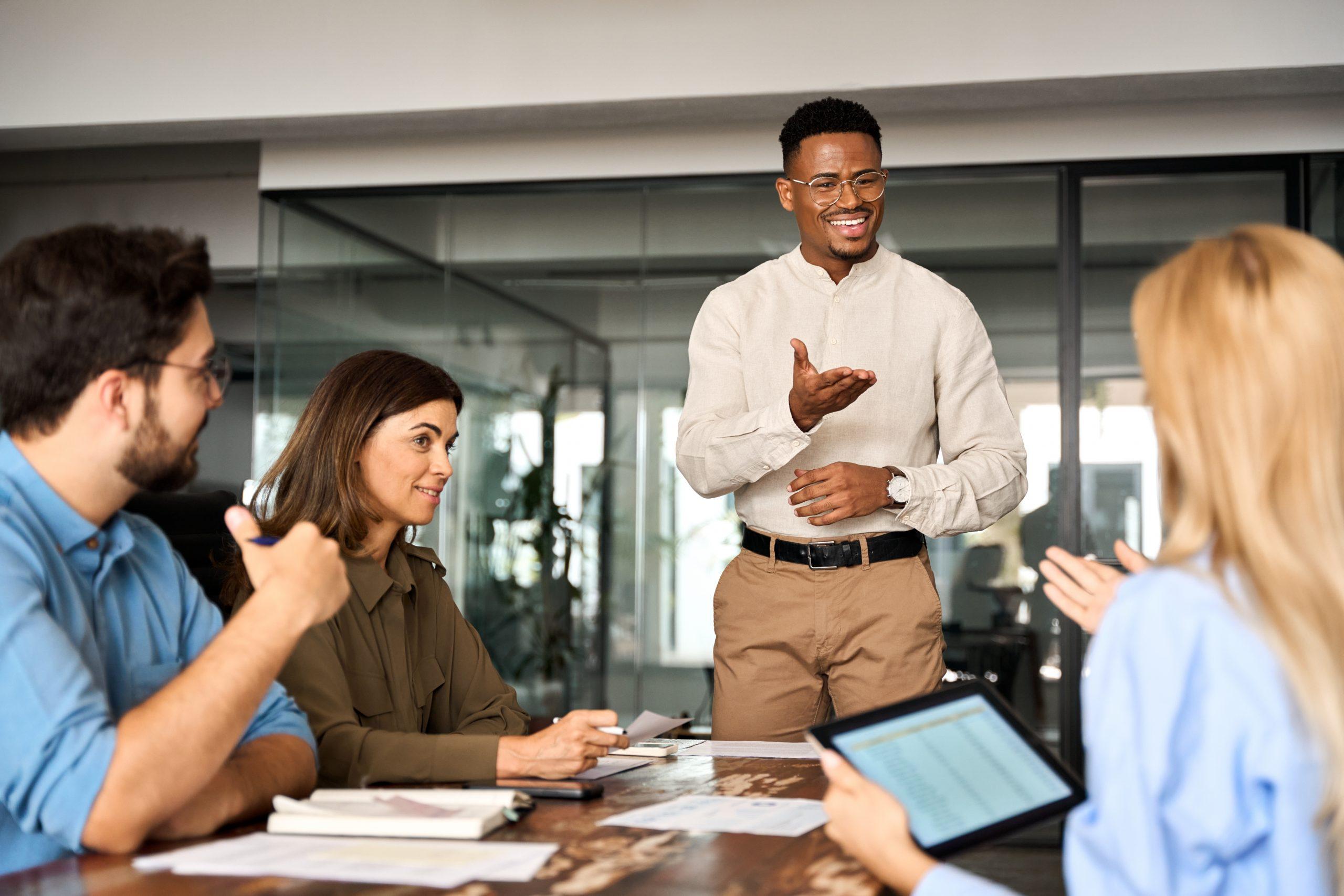 A man confidently presents to colleagues in a modern office as they listen and engage.