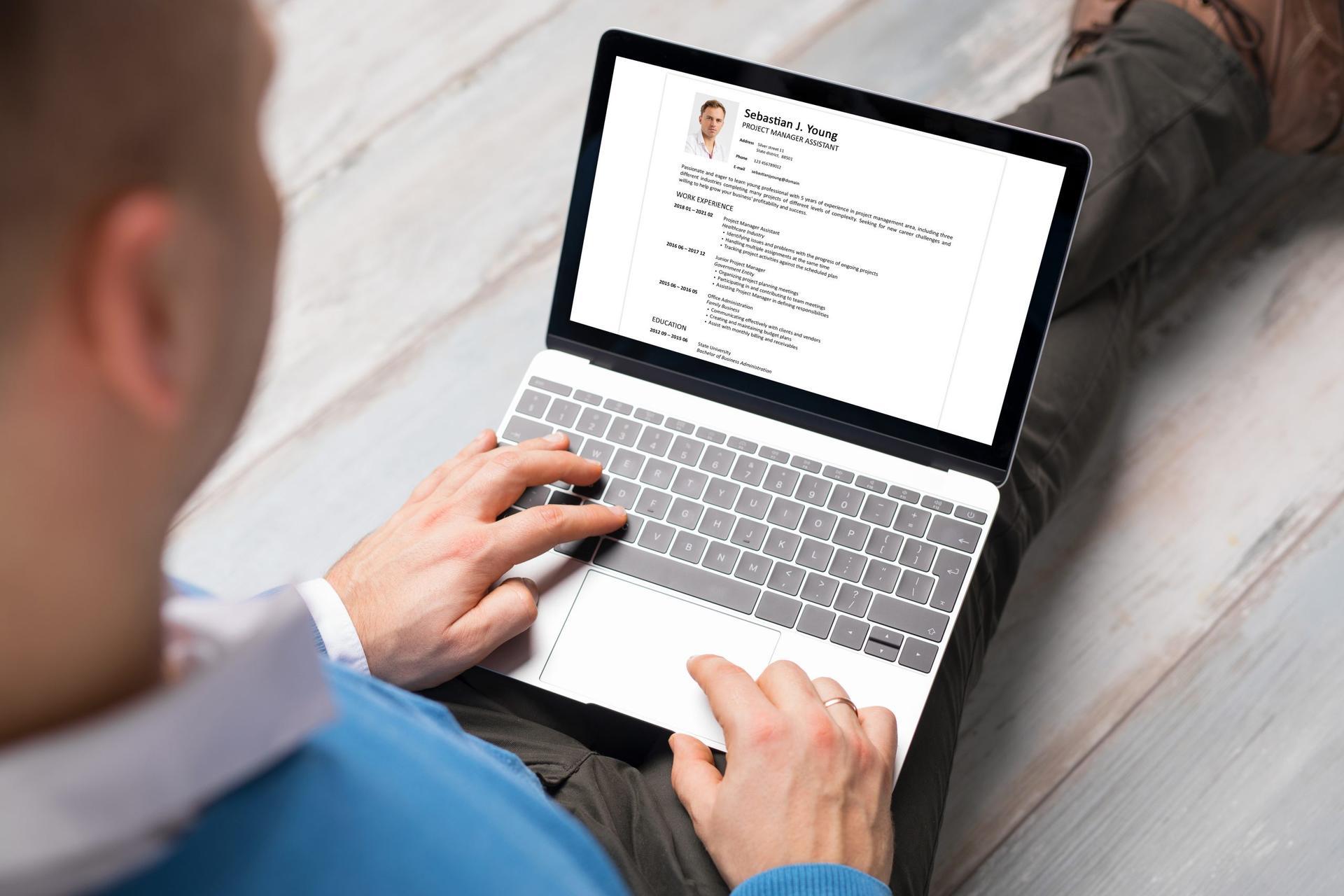 Over-the-shoulder view of man using laptop to review and update resume while sitting on floor.